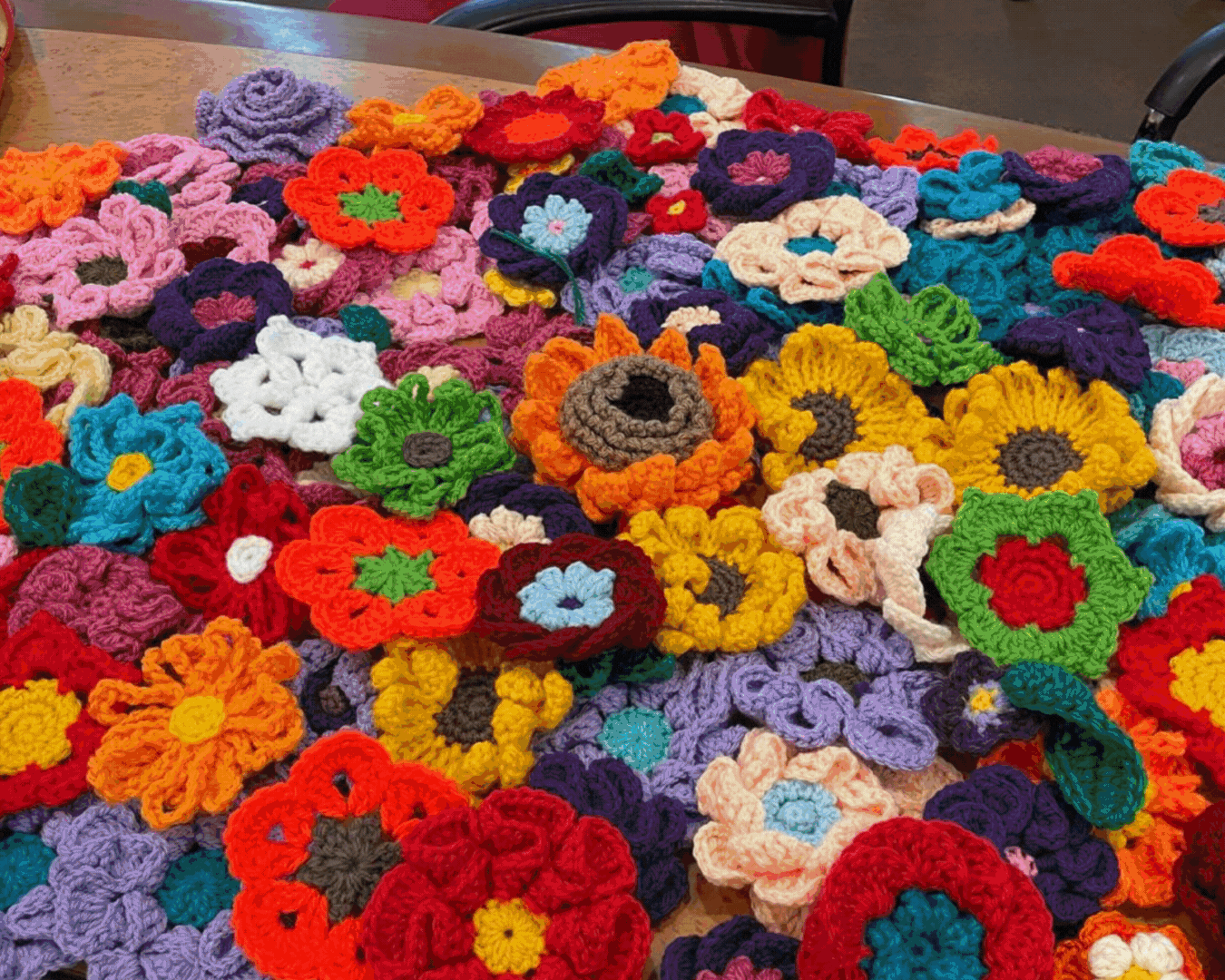 Dozens of colorful crocheted yarn flowers in red, yellow, purple, orange, and teal spread across a wooden table.